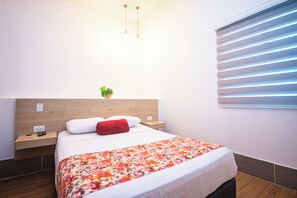 A hotel room with a wooden headboard, a bed with a floral bedspread, a red pillow, a nightstand with a remote control, and a small plant on the headboard. - HOTEL BOUTIQUE POMA ROSA (Medellin)