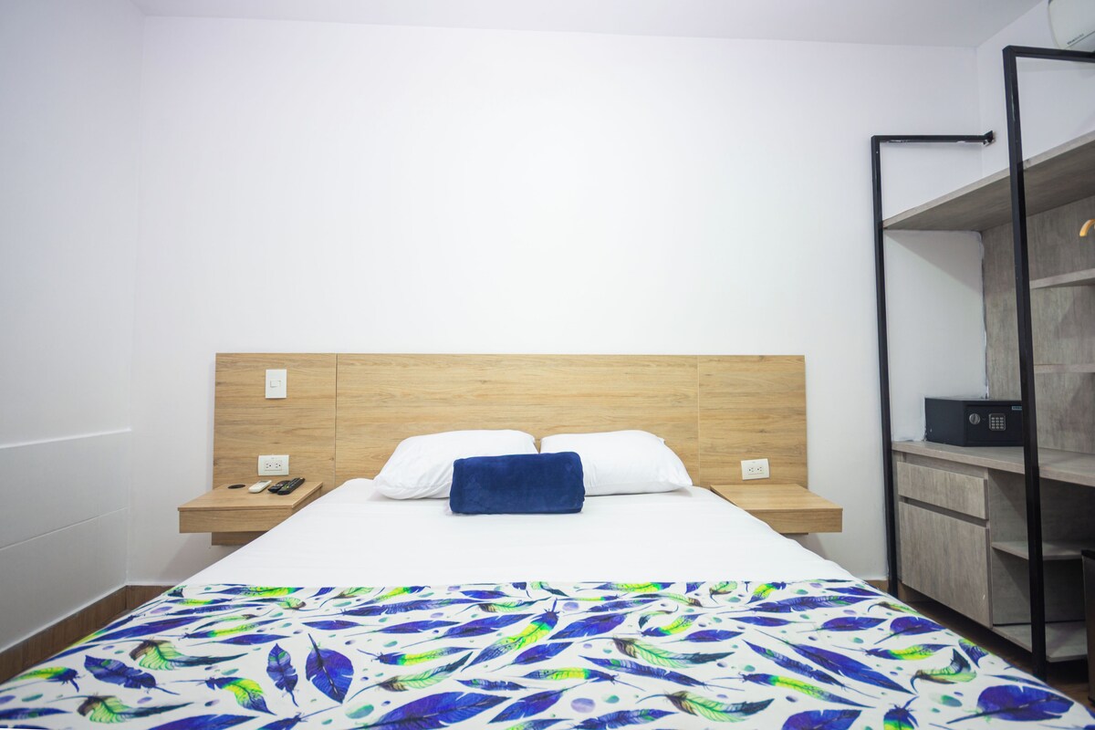 A hotel room with a wooden headboard, a bed with white linens, a blue throw blanket, a bedside table with a phone and remote controls, and a built-in shelving unit.