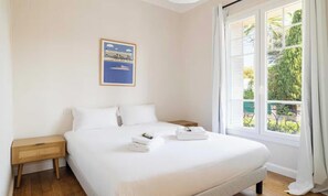 A hotel room with a large bed, a nightstand, a framed picture on the wall, and a window with a view of greenery. - DISCOVER THE CHARM OF THE FRENCH RIVIERA IN THE HEART OF CANNES. (Cannes)