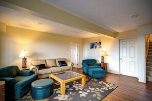 Ein Wohnzimmer mit einer Sofagarnitur, einem Couchtisch und zwei Sesseln. Im Hintergrund sind eine Tür und eine Treppe zu sehen.