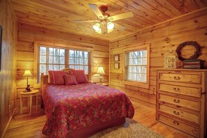 Une chambre dans une cabane en bois, avec un lit, une commode, deux fenêtres et un ventilateur de plafond.