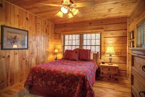 A wooden cabin bedroom with a bed, two bedside lamps, a ceiling fan, and a window. - Arbor Den Log Cabin 3 Bedroom, 3 Bath, Pet friendly (Blowing Rock)