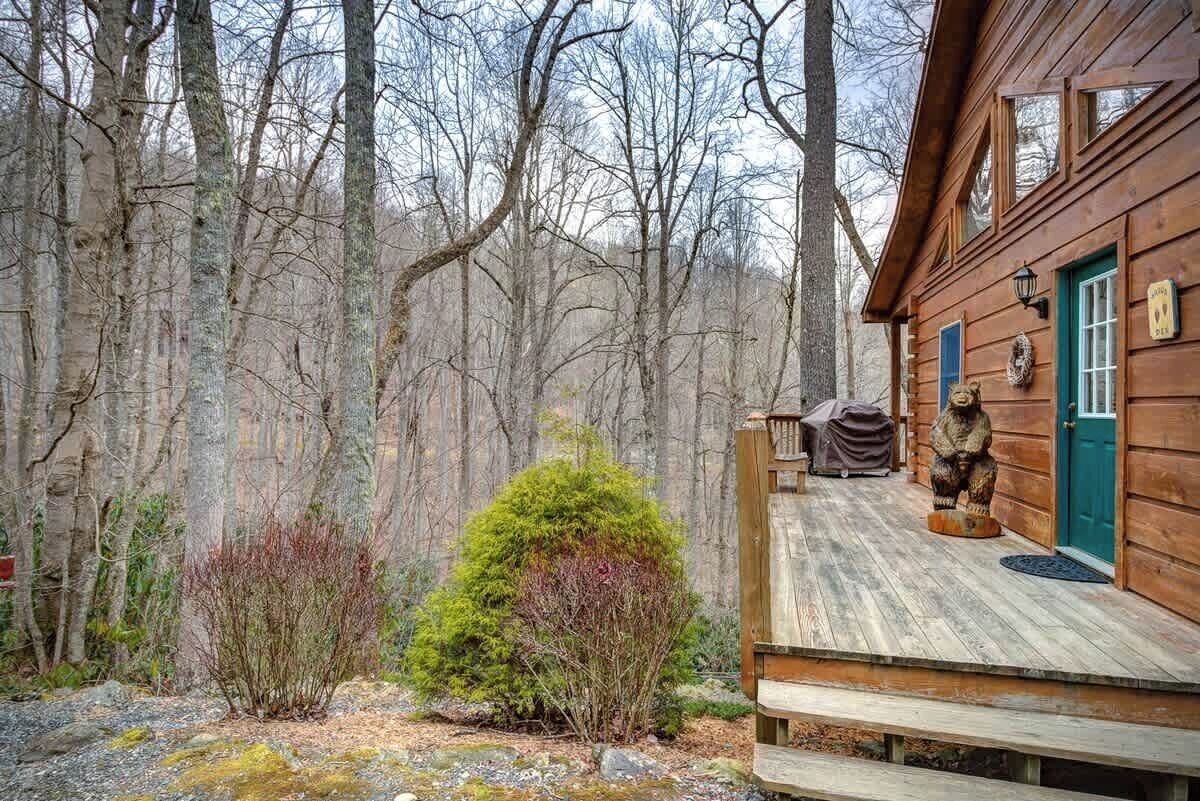 A wooden cabin with a porch, surrounded by trees and a small bush.