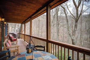 A covered outdoor deck with wooden railings, a table with chairs, and a view of a forested area. - Arbor Den Log Cabin 3 Bedroom, 3 Bath, Pet friendly (Blowing Rock)