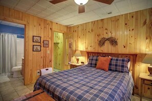 Une pièce aux murs lambrissés, comprenant un lit, une table de chevet, une lampe fixée au mur et une porte donnant sur une autre pièce.