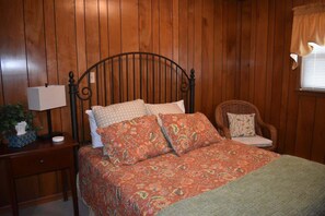 A wooden-paneled room with a bed, a nightstand, a lamp, a wicker chair, and a window with a curtain. - Minute to Main - Steps to Main St Blowing Rock (Blowing Rock)