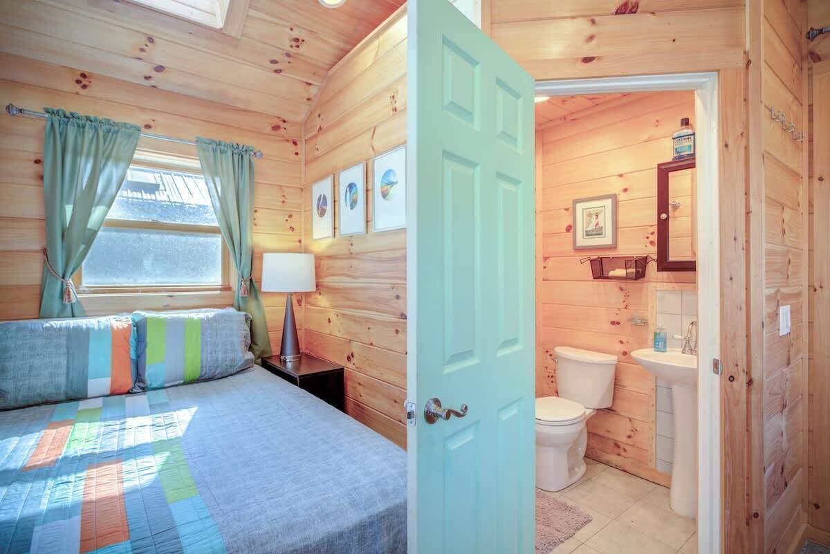 A wooden cabin room with a bed, a window with curtains, a door leading to a bathroom with a toilet and sink.