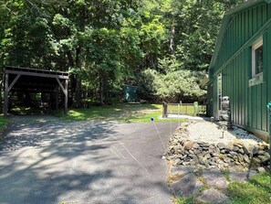 A paved driveway leading to a covered parking area, surrounded by trees and a green building with a stone feature.