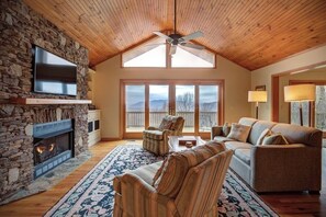 A cozy living room with a stone fireplace, a television, and a large window overlooking a mountainous landscape. - Fair Haven - Great App Ski Mtn Location! (Blowing Rock)