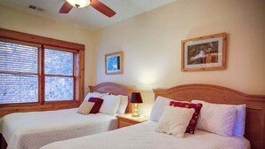 Quarto de hotel com duas camas, um ventilador de teto, uma janela com persianas, um quadro na parede e uma mesa de cabeceira com abajur.