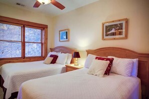 Ein Hotelzimmer mit zwei Betten, einem Deckenventilator, einem Fenster mit Jalousien, einem gerahmten Bild an der Wand und einem Nachttisch mit Lampe.