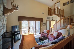 A living room with a stone wall, a mounted deer head, a television, a fireplace, a staircase, and a large window. - One Level Entry 3 bed 3 bath condo at Chetola (Blowing Rock)