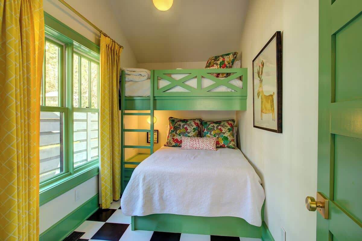 A room with a bunk bed, a window with curtains, a framed picture on the wall, and a door with a brass handle.
