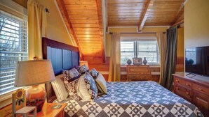 A bedroom with a wooden ceiling, a bed with patterned bedding, a nightstand with a lamp, and a dresser with a television. - 3 Bedroom 3 Bath Log Cabin just outside of Blowing (Blowing Rock)