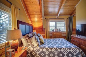 A bedroom with a wooden ceiling, a bed with patterned bedding, a nightstand with a lamp, and a dresser with a television. - 3 Bedroom 3 Bath Log Cabin just outside of Blowing (Blowing Rock)