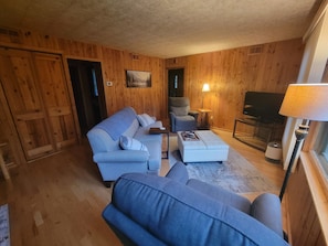A cozy living room with a sofa, ottoman, and armchair, wooden walls, a television, and a lamp. - Cozy 3-bedroom cabin in serene Park Rapids (Park Rapids)