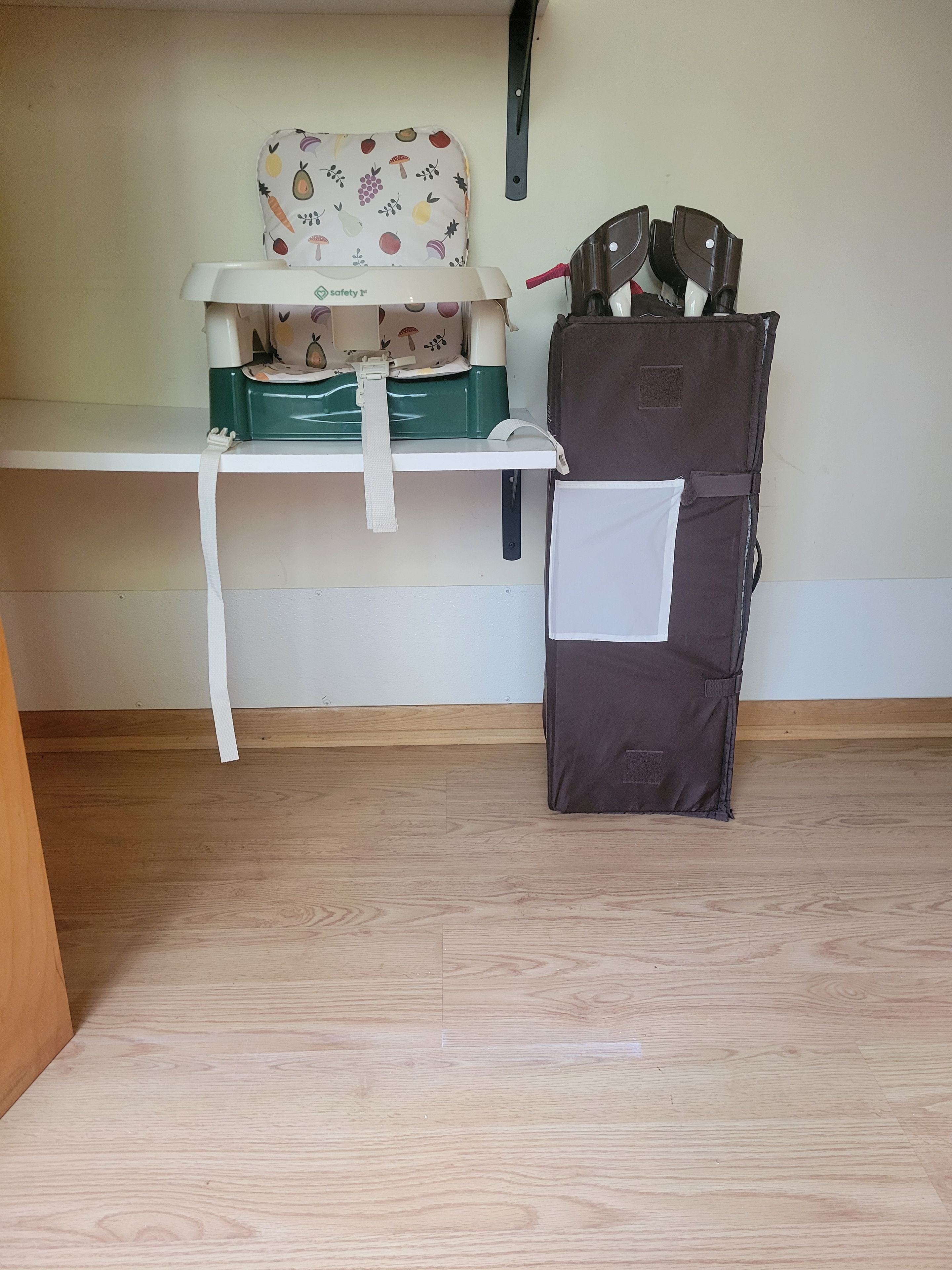 A room with a wooden floor, a white shelf, a green and white baby changing station, and a brown storage bag.
