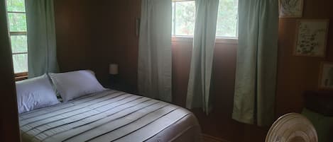 A bedroom with a bed, a nightstand, a wall-mounted fan, and two windows with curtains.