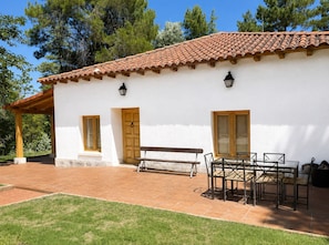 A white building with a tiled roof, wooden door, and windows, a bench, and a table with chairs outside.