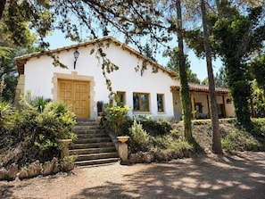 A white building with a wooden door and windows, surrounded by greenery and a stone staircase leading to the entrance.
