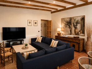 A living room with a dark blue sofa, a flat-screen TV, a wooden coffee table, wicker chairs, and a painting on the wall.