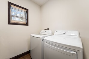 A laundry room with two white front-loading washing machines and a window with blinds. - Maplecrest Retreat | Firepit • Hot Tub • Game Room (Sevierville)