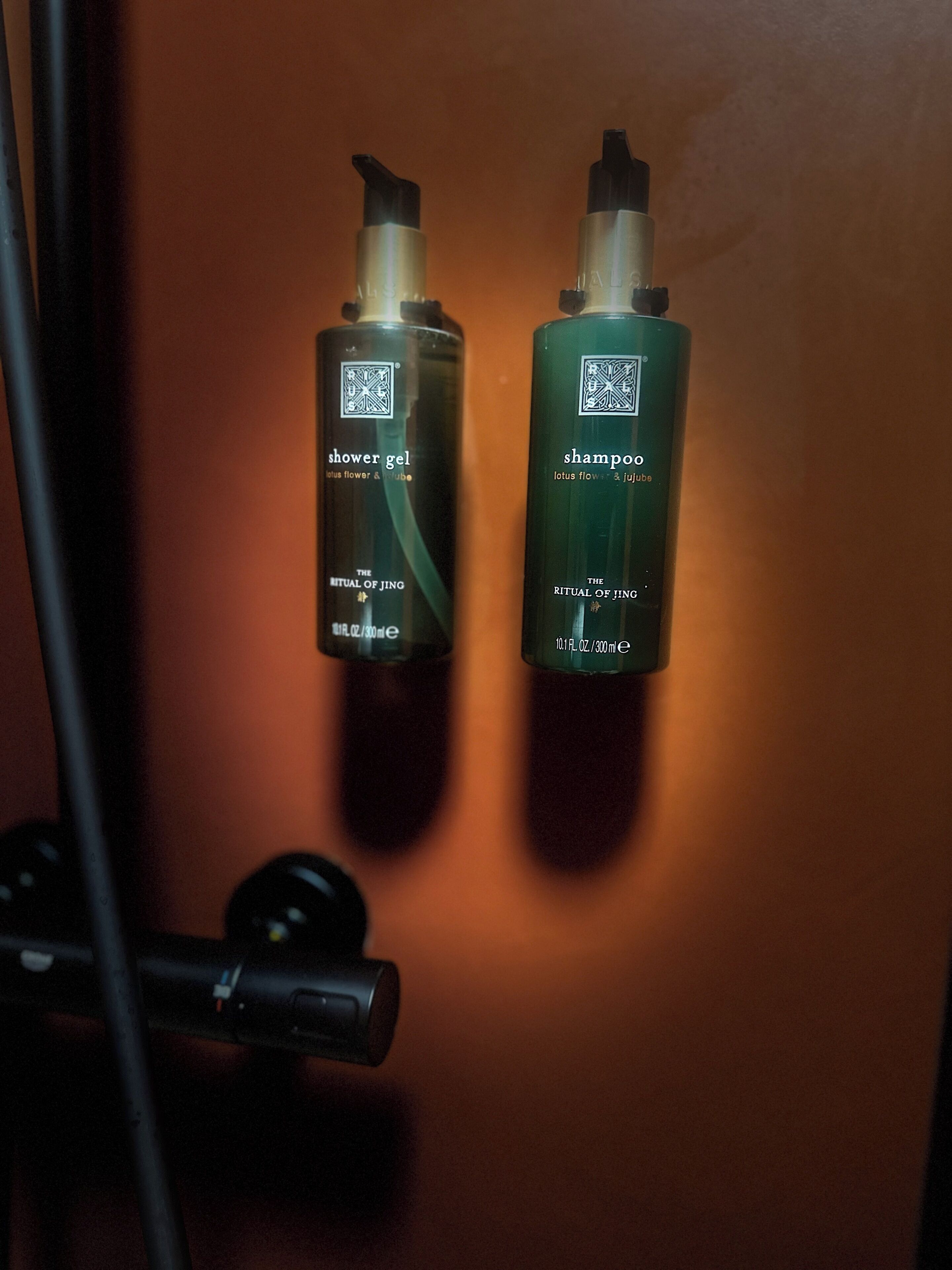 Two bottles of The Ritual of Jingo shower gel and shampoo, with a showerhead in the foreground.