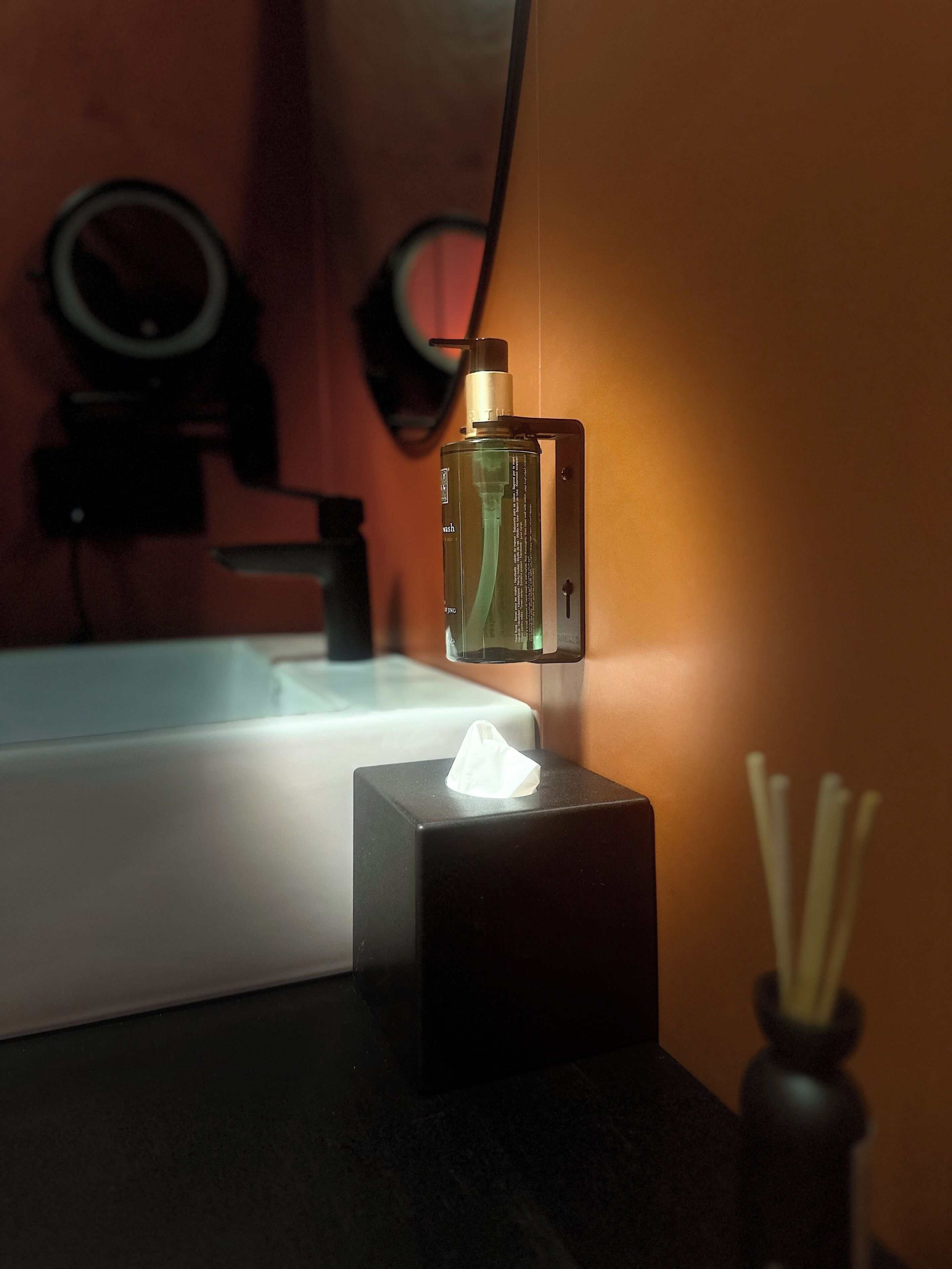 A bathroom with a hand soap dispenser mounted on the wall, a tissue box, and a reed diffuser.