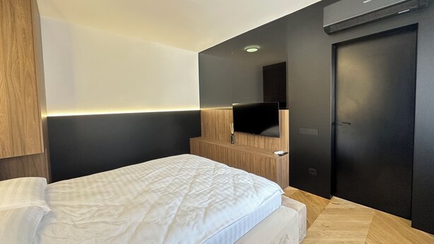 A modern hotel room with a bed, a flat-screen TV mounted on the wall, a wooden headboard, and a door.