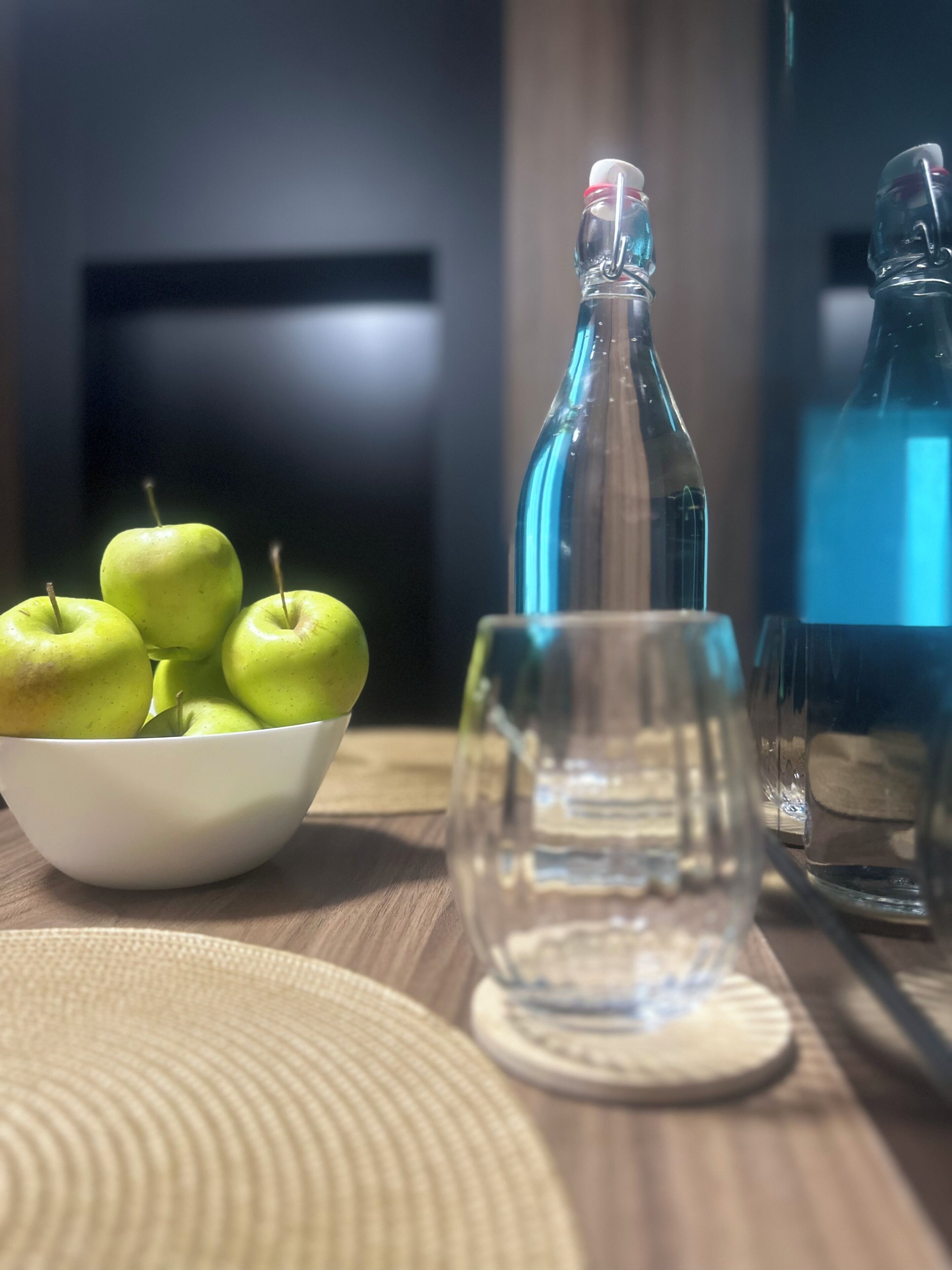 A table with a bowl of green apples, a glass bottle, and a glass.