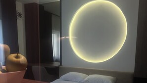 A hotel room with a bed, a round wall light, a bowl of fruit, and a mirror.