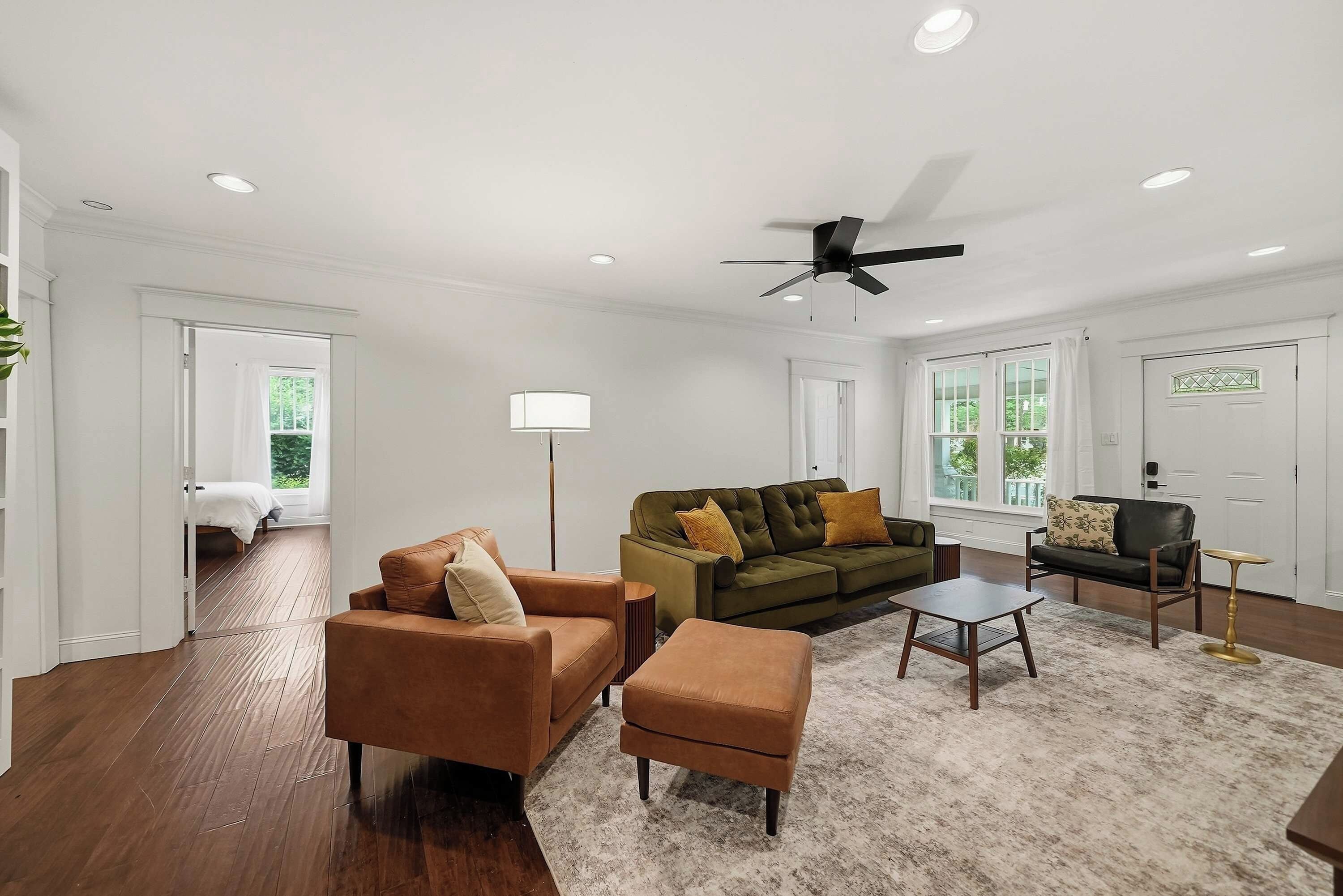 A spacious living room with a ceiling fan, a sofa set, a coffee table, and a view of a bedroom through an open door.