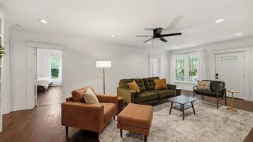 A spacious living room with a ceiling fan, a sofa set, a coffee table, and a view of a bedroom through an open door.