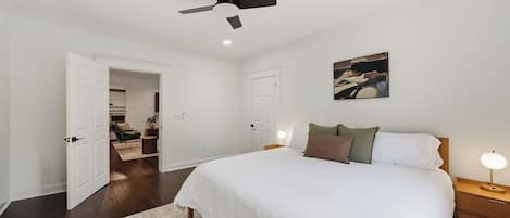 Un dormitorio con cama, mesitas de noche, ventilador de techo, un cuadro y un pasillo que conduce a otra habitación.