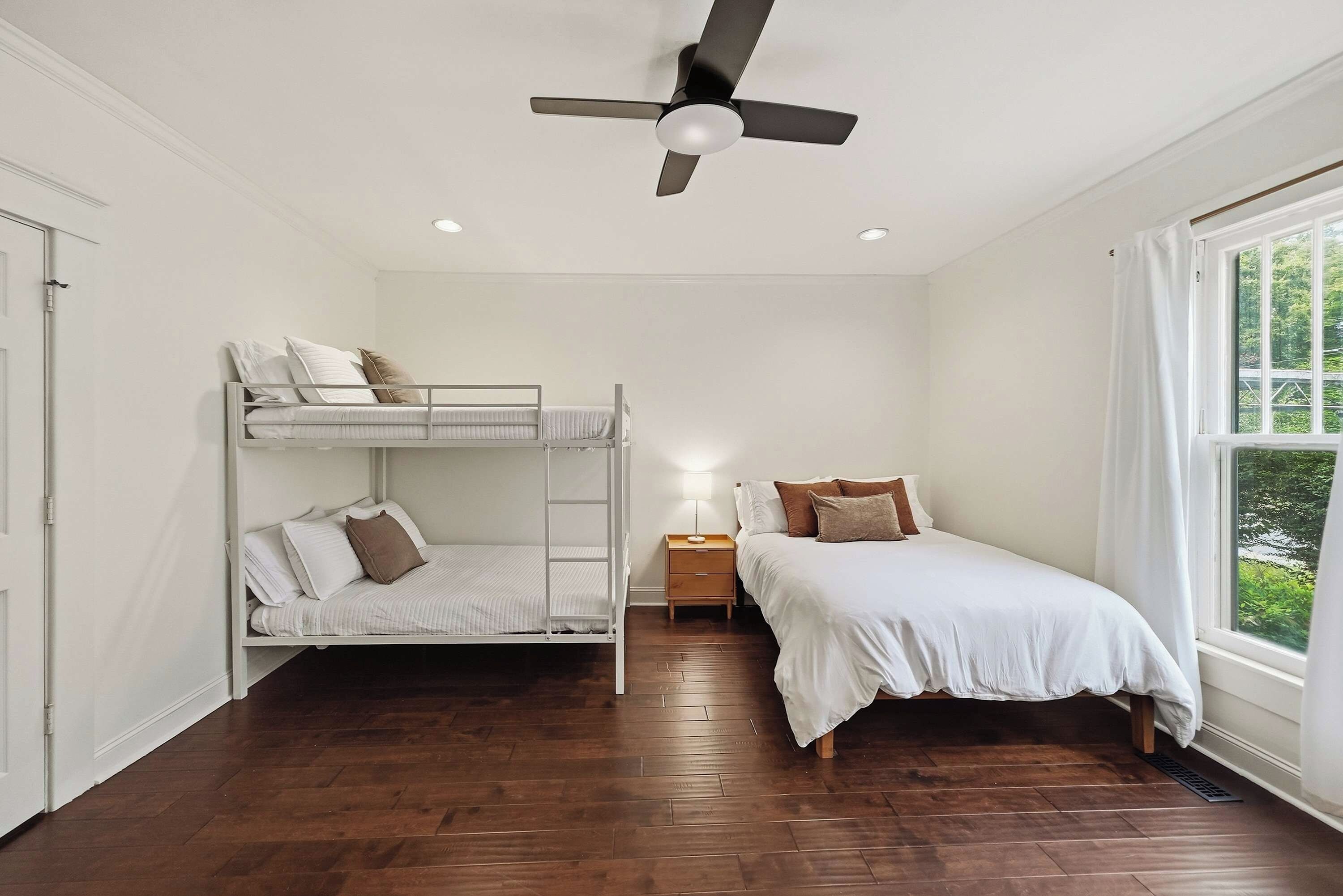 A room with a bunk bed, a single bed, a nightstand, a ceiling fan, and a window with a view of greenery.