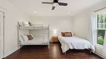 A room with a bunk bed, a single bed, a nightstand, a ceiling fan, and a window with a view of greenery.