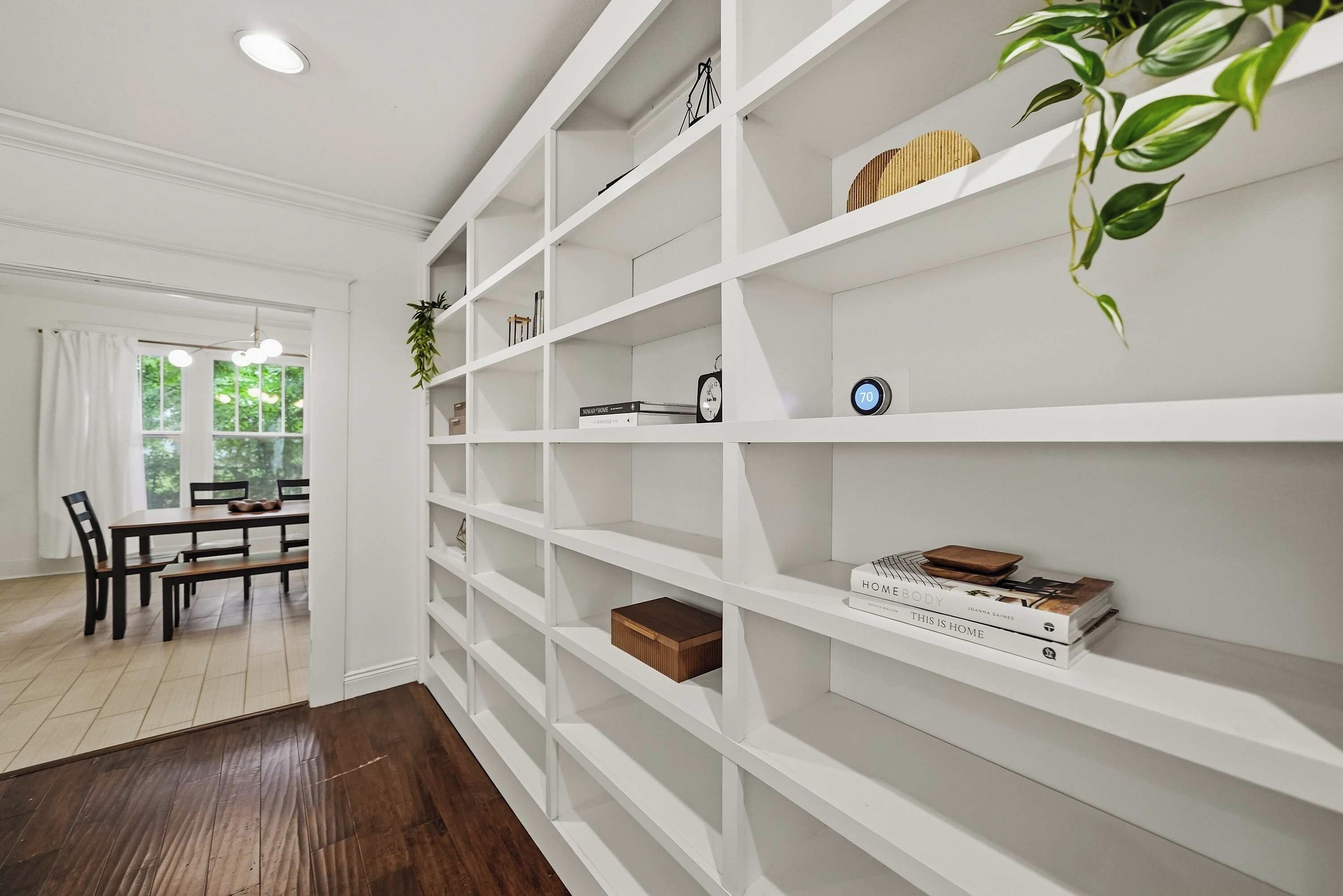 Una habitación con una estantería blanca, un comedor con mesa y sillas, y una planta.
