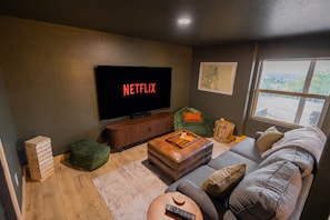 A living room with a television displaying 'NETFLIX,' a sofa, a coffee table, a bean bag, and a window with a view. - NEW! A-Frame, Hot Tub, MTN Views, Game & Movie Room (Divide)