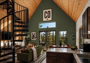 A spacious loft with a high ceiling, a spiral staircase, a large window, and a kitchen area. - Designer A-Frame, Hot Tub, Mountain Views, Game Room (Divide)
