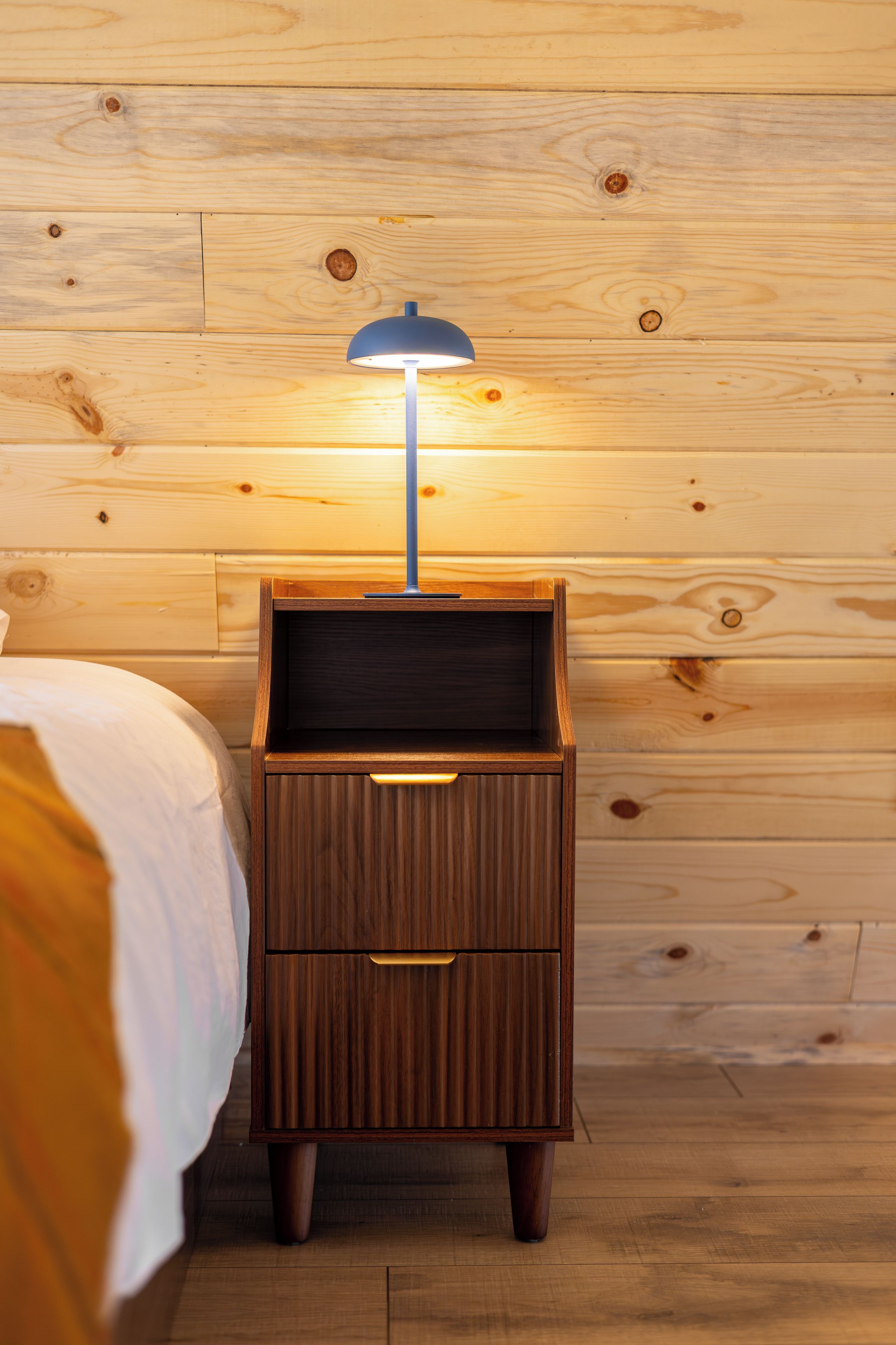 Une table de chevet en bois avec une lampe, un tiroir et un lit recouvert d’une couvre-lit blanche.