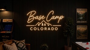 A cozy room with a neon sign reading 'Base Camp Colorado,' a Pac-Man arcade machine, a leather couch, a coffee table with books, and a poster of Colorado's mountains. - Designer A-Frame, Hot Tub, Mountain Views, Game Room (Divide)