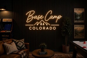 A cozy room with a neon sign reading 'Base Camp Colorado,' a Pac-Man arcade machine, a leather couch, a coffee table with books, and a poster of Colorado's mountains. - Designer A-Frame, Hot Tub, Mountain Views, Game Room (Divide)
