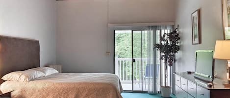 A bedroom with two beds, a dresser, a plant, and a balcony with a view of greenery.