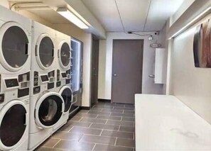 Un lavadero con varias lavadoras, piso de baldosas y una puerta que da a otra habitación.