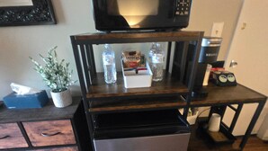 Eine kompakte Küchenzeile mit Mikrowelle, Kühlschrank und einer kleinen Ablage mit Gegenständen wie Wasserflaschen und einem Snackriegel.