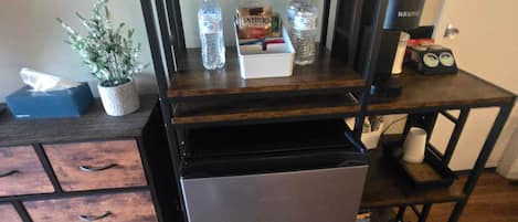 A compact kitchenette with a microwave, refrigerator, and a small shelf with items like water bottles and a box of snacks.