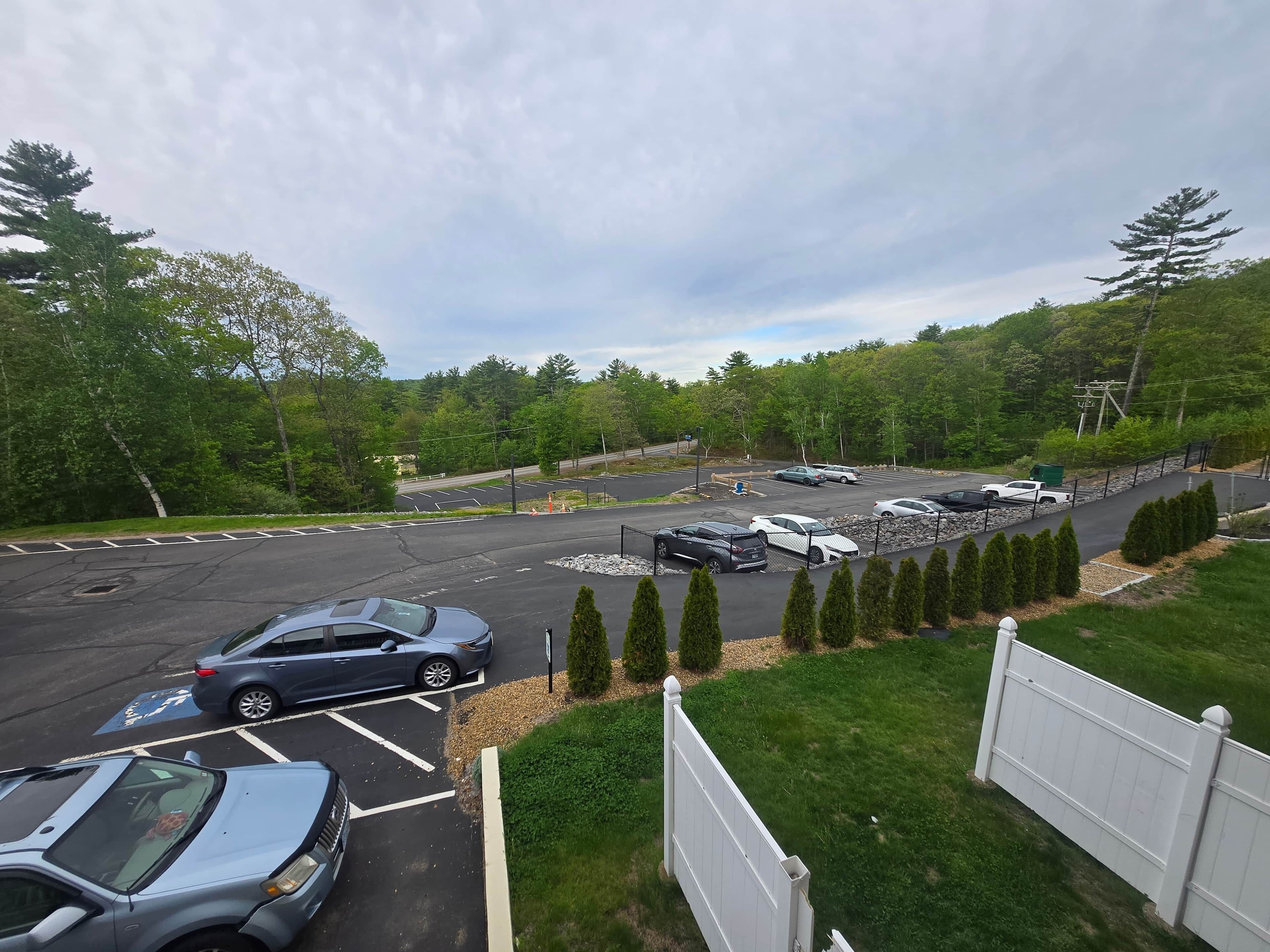 Un parking rempli de voitures, une clôture blanche et des arbres verts en arrière-plan.