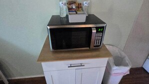 Eine Mikrowelle auf einem Schrank mit einem kleinen Regal darüber, auf dem sich eine Wasserflasche, eine Tüte mit Snacks und eine Taschentuchbox befinden.