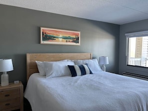 Cama bem arrumada com roupa de cama branca e almofada listrada, cabeceira de madeira, mesa de cabeceira com abajur e um quadro de uma paisagem na parede.