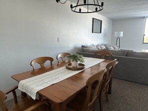 Een eetgedeelte met een houten tafel, stoelen, een geweven placemat en een kamerplant. Op de achtergrond staat een grijze bank en een vloerlamp.